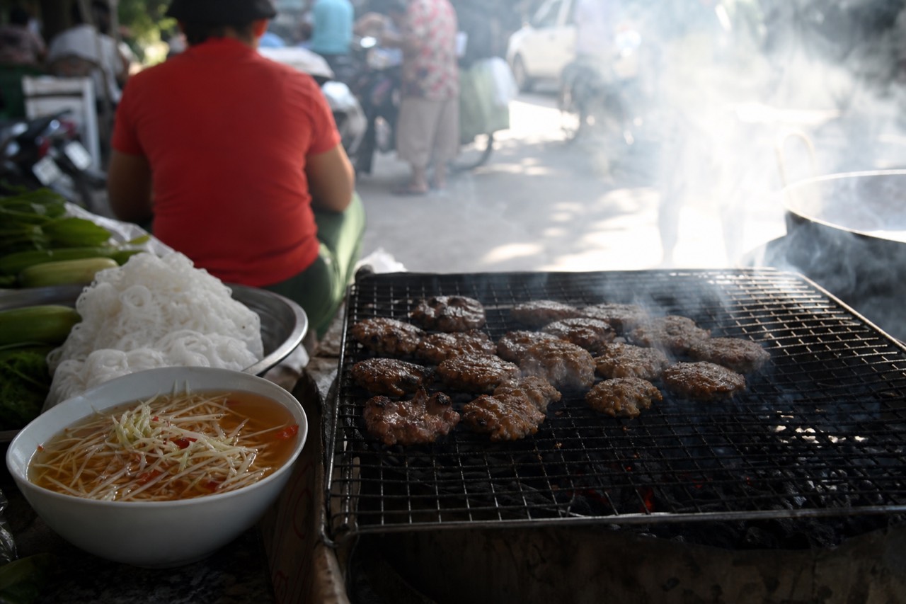 Grilled pork patties over a charcoal fire beside bowls of bun cha dipping sauce in Hanoi
