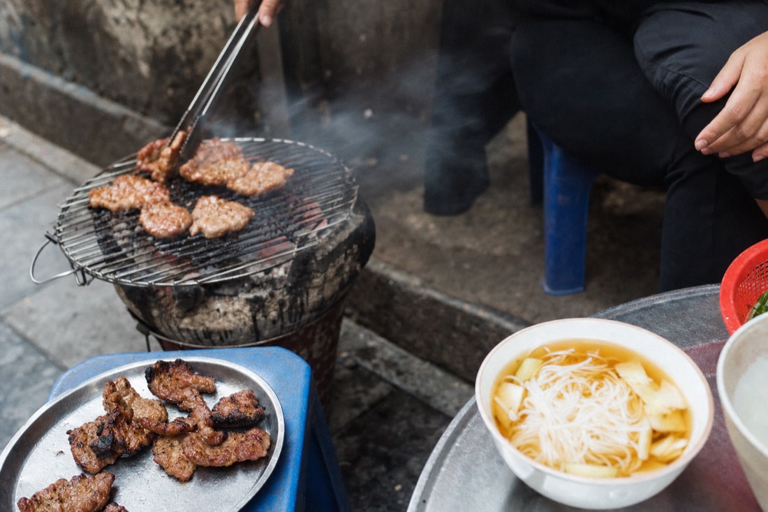 Bun cha grilling over a charcoal brazier on a Hanoi pavement, smoke drifting past a bowl of dipping broth