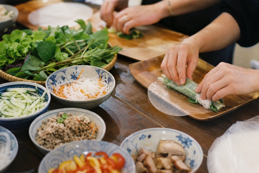 Hands rolling fresh spring rolls at a Hanoi cooking class table with herbs and fillings