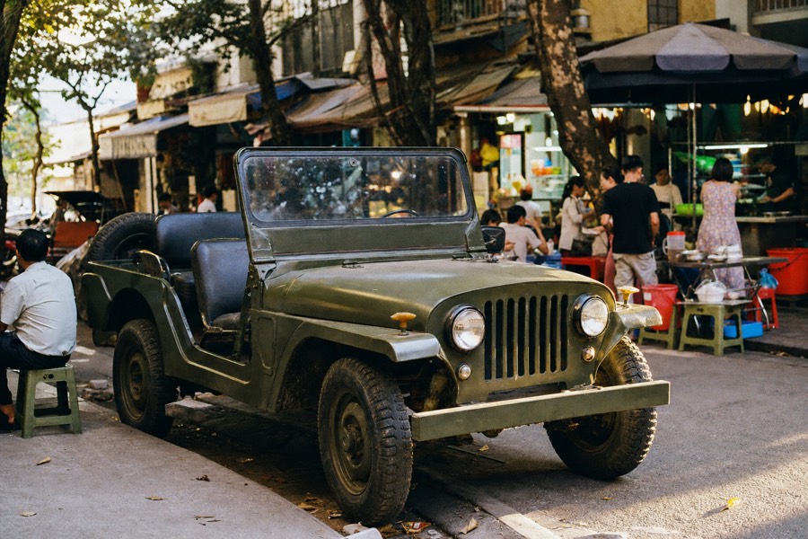 A vintage open-top army jeep parked on a Hanoi street corner near food stalls