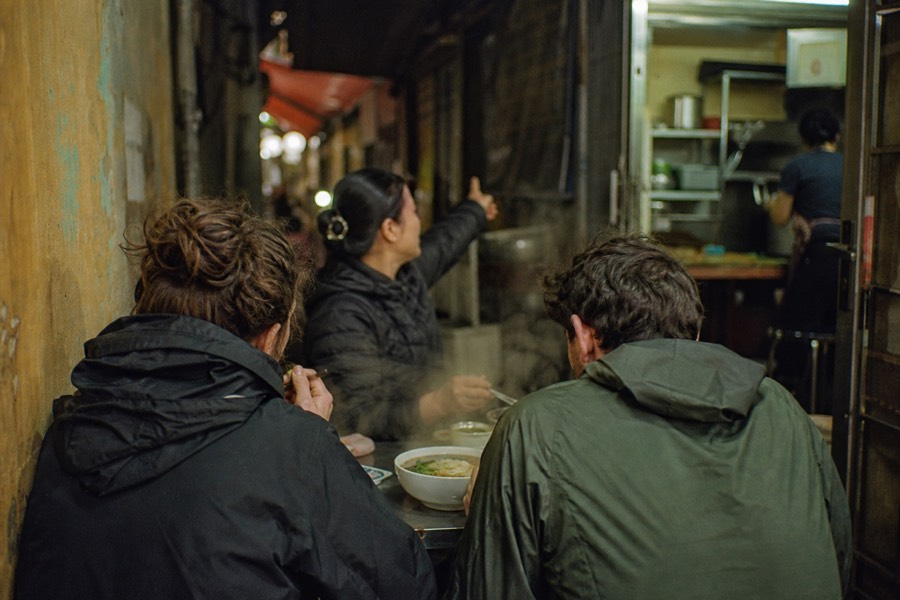 Two travelers eating noodle soup at a quiet Hanoi alley table on a private food walk